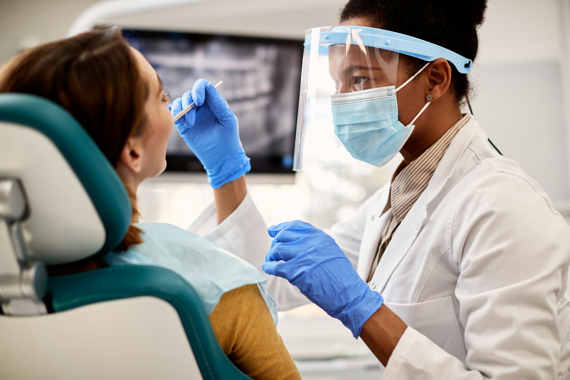 Female dentist examining a woman's teeth while wearing a protective visor for safety and hygiene.